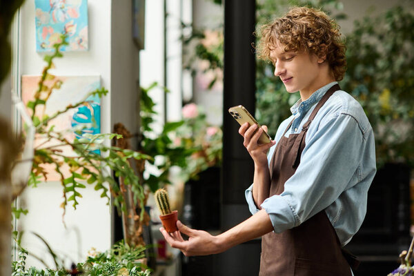 A young man in an apron explores a small cactus while chatting on his phone in a plant shop.