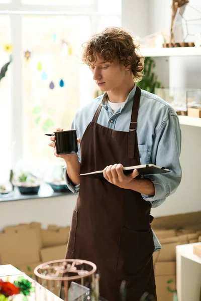 Önlüklü genç bir adam kahveyi yudumluyor ve yaratıcı bir mekanda eskizler çiziyor..
