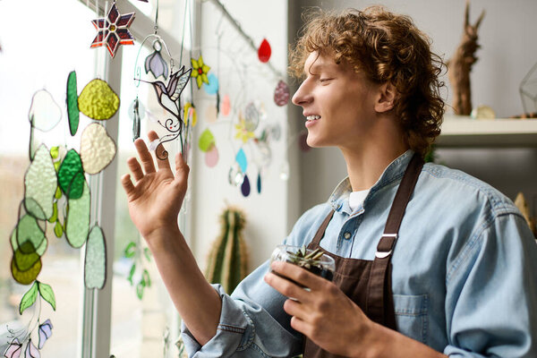 Young man admires colorful stained glass creations while holding a small plant in a bright studio.