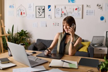 Dövmeleri olan genç bir kadın, rahat bir evde sanat eseri yaratıp telefonuyla sohbet etmekle meşgul..