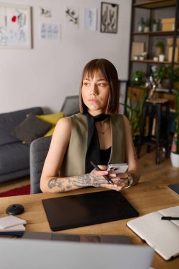 Genç yaratıcı kadın evinde, elinde bir telefon ve kalemle düşünceli bir şekilde oturuyor..