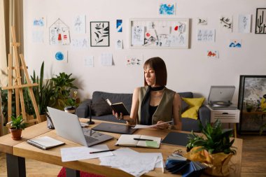 Genç bir kadın sanatsal çalışma alanı ile çevriliyken defteriyle meşgul oluyor..