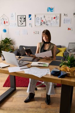 Genç bir kadın evindeki rahat ofisinde yaratıcı materyallerle çevrili iken notlarını yansıtıyor..