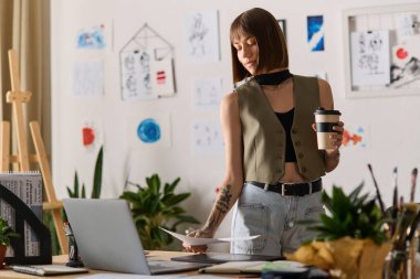 Genç bir kadın evde, bitkiler ve yaratıcılıkla çevrili sanat eserlerini incelerken kahvesini yudumluyor..