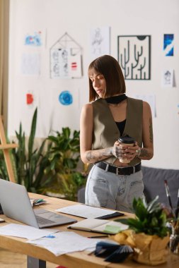 Genç bir kadın düşünceli bir şekilde masasında duruyor, elinde kahveyle, yaratıcı alanında..