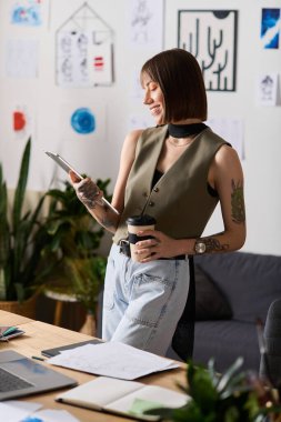 Evde yaratıcı işler yapan genç bir kadın elinde kahve fincanı tutarken tablet kullanıyor..