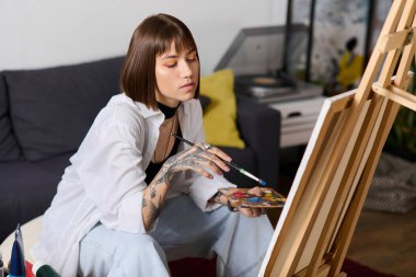 Dövmeli genç bir kadın şık ev ortamında resim yapmaya odaklanıyor..