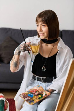 Genç, yaratıcı bir kadın evinde dinleniyor, çayını yudumluyor ve elinde renklerle dolu bir boya paleti tutuyor..