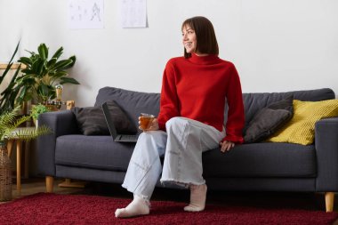 Genç bir kadın çayını yudumlarken ve parlak bir odada laptopuyla çalışırken kanepede dinleniyor.