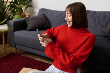 Samimi kırmızı kazak giymiş genç bir kadın evde akıllı telefonunu kullanırken gülümsüyor..