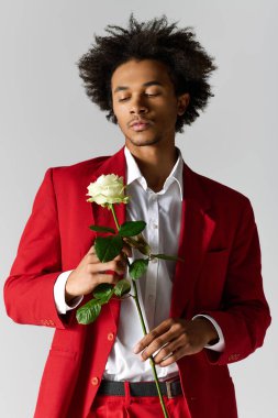 Şık kırmızı takım elbiseli genç bir adam nazikçe beyaz bir gül tutuyor, zarafet saçıyor ve dengede duruyor..