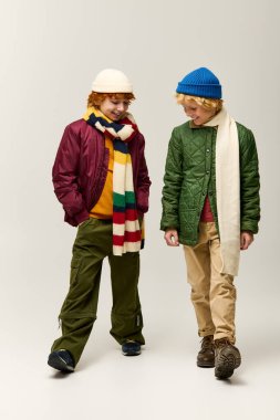 İki neşeli çocuk birlikte yürür, şık kış kıyafetlerini oyuncu bir şekilde sergilerler..