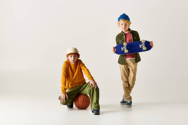 İki şık çocuk, parlak bir stüdyoda bir basketbol topu ve kaykayla neşeli titreşimler paylaşıyor..