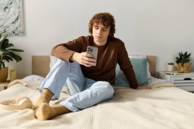 Kıvırcık saçlı bir genç rahat bir şekilde yatağında oturur, rahat yatak odasında akıllı telefonunu kontrol eder..