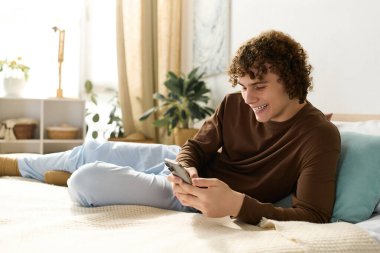 Genç bir kız rahat yatak odasında, telefonunu mutlu bir şekilde kullanıyor ve yatakta dinleniyor..