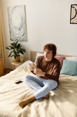 Kıvırcık saçlı bir genç, rahat bir yatak odasında yatağında, kulaklıklarla meşgul.