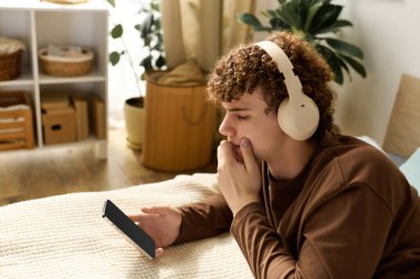 Kıvırcık saçlı ergen çocuk rahat bir odada yatağında uzanırken müzik dinliyor..