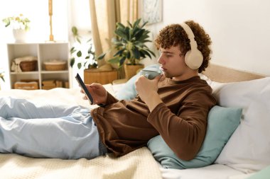 Ergen çocuk yatağında vakit geçirmekten zevk alıyor, akıllı telefonunu kullanırken bir yudum içki içiyor..