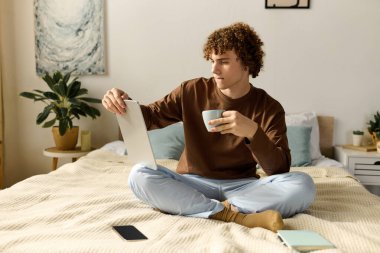 Kıvırcık saçlı ergen çocuk yatağında bacak bacak üstüne atmış, çayını yudumluyor ve tablet kullanıyor..