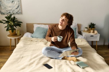 Kıvırcık ergen bir çocuk sıcak bir odada tabletini içerken kahvesini yudumluyor..