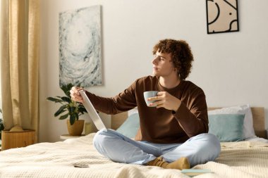 Kıvırcık saçlı bir genç elinde bir bardakla yatakta rahatlar ve tablete bakar..