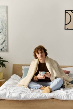 Kıvırcık saçlı bir genç, şık yatak odasında çapraz bacaklı otururken sıcak bir içkiden zevk alır..