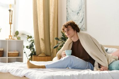 Kıvırcık saçlı bir genç yatağa oturur, battaniyeye sarılır ve içkisini yudumlar..