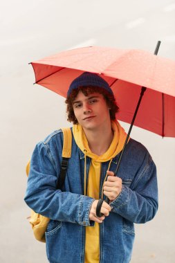Genç bir çocuk, bir sonbahar günü elinde kırmızı bir şemsiye, kot pantolon giymiş, dışarıda duruyor..