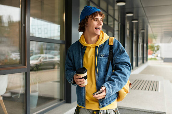 A cheerful teenager wearing a blue beanie and denim jacket stands outside in autumn, sipping coffee.
