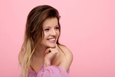 Pembe makyajlı genç bir kadın stüdyoda organza bir bluzla gülümsüyor..