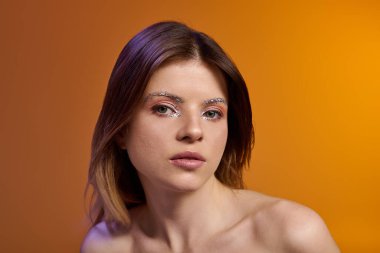 Çekici genç bir kadın renkli bir stüdyoda göz makyajı ve çıplak omuzlarıyla poz veriyor..