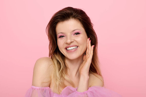 Молодая женщина с розовым макияжем ярко улыбается в студии, демонстрируя свою модную блузку.