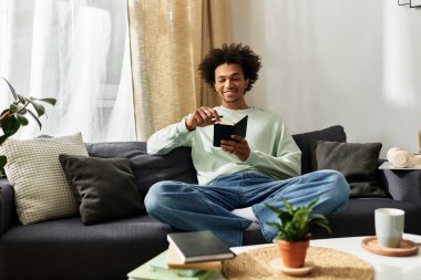 Genç bir Afrikalı Amerikalı adam rahat bir kanepede rahatlar, cihazını kullanırken kendini evinde hisseder..