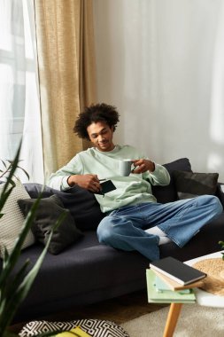 Genç Afrikalı Amerikalı adam kanepede kitap okurken evinde sıcak bir içecekle rahatlıyor..