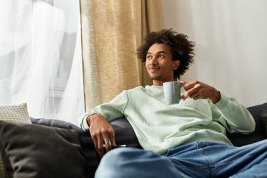 Genç adam evinde rahatça oturur, kahvesini yudumlar ve huzurlu bir öğleden sonranın tadını çıkarır..