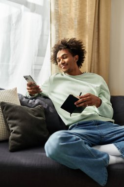 Yakışıklı genç adam rahat bir kanepede dinleniyor, modern bir yaşam alanında elinde bir defter ve telefon tutuyor..