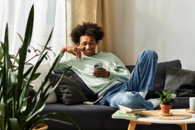 Yakışıklı bir Afrikalı genç adam rahat bir kanepede rahatlar, telefonunu kullanırken gülümser..