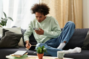 Genç adam modern oturma odasında dinleniyor, notlar yazarken telefonuna odaklanıyor..