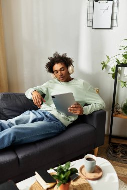 Yakışıklı genç bir adam kanepede dinleniyor, müzikten zevk alıyor ve tabletini evde karıştırıyor..