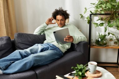 Yakışıklı genç bir adam rahat bir yaşam alanındaki kanepede uzanır ve tabletine odaklanır..