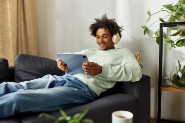 Yakışıklı genç bir adam kanepede uzanıyor, rahat bir yaşam alanında tabletinden müziğin tadını çıkarıyor..