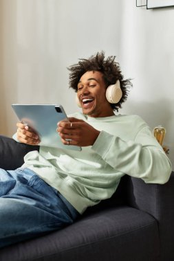 Neşeli bir Afrikalı genç Amerikalı adam tabletine gülerken rahat bir kanepede dinleniyor..