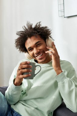 Neşeli genç adam modern evinde dinleniyor, kahvesini yudumluyor ve kulaklıkla müzik dinliyor..