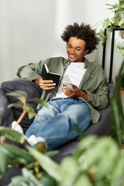 Genç bir Afrikalı Amerikalı adam evde rahatlıyor, kitap okurken gülümsüyor ve telefonunu kullanıyor..