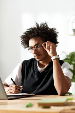 Kıvırcık saçlı genç adam bir masada oturuyor, dizüstü bilgisayar kullanırken notlar yazıyor..