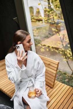 Beyaz bornozlu güzel bir kadın kaplıcada rahatlar, bir yudum içer ve manzaranın tadını çıkarır..