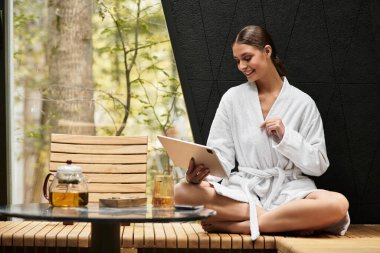 Yumuşak beyaz bornozlu genç bir kadın huzur içinde oturup spa deneyiminin tadını çıkarıyor..