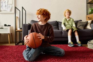 İki neşeli çocuk evde eğlenceli bir anı paylaşıyor, biri halının üzerinde otururken elinde basketbol topu tutuyor..