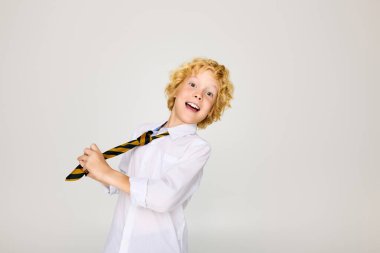 Dalgalı sarı saçlı genç bir çocuk üniforma giyerken çizgili kravatını heyecanla düzeltiyor..