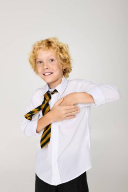 Okul üniformalı neşeli sarışın bir çocuk, dalgalı saçlarını sergileyerek stüdyoda şakacı bir poz veriyor..